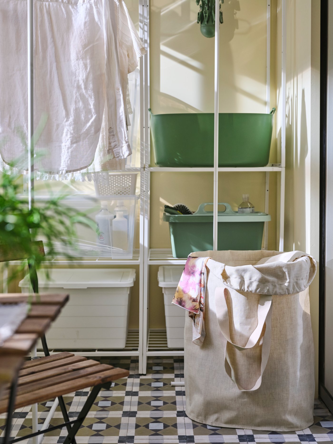A beige PURRPINGLA laundry bag with handles is on the floor of an enclosed balcony beside a shelving unit and drying rack.