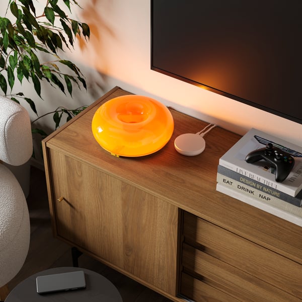 A VARMBLIXT LED table/wall lamp sits near a DIRIGERA hub and some books on a RÅDMANSJÖ TV bench underneath a wall-mounted TV.