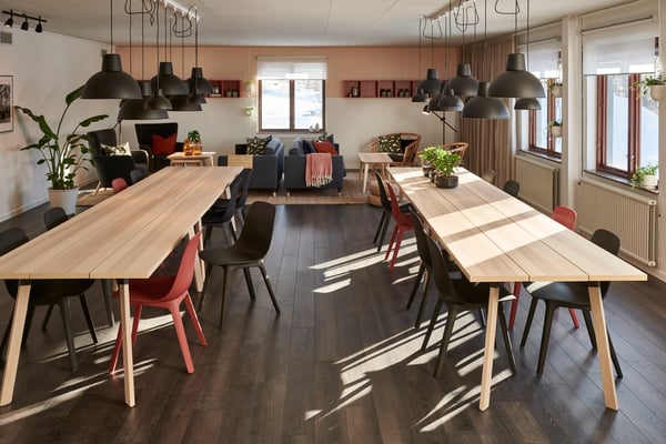 Efter-bild av matsalen på hur den ser ut efter renoveringen. Rummet har nu mörkt trägolv och långa bord i ljust trä och svarta och röda stolar.