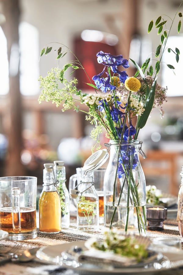 Wildblumenstrauss in Glasvase, daneben Gläser und Flaschen auf gedecktem Tisch.