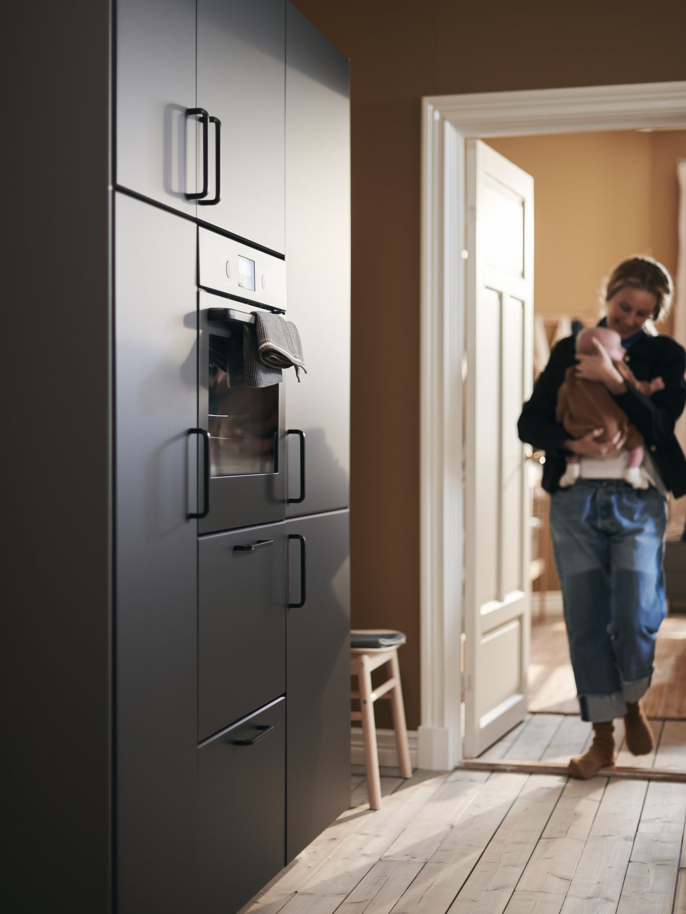 A mother cradles her baby as she enters the kitchen, approaching a wall of cabinets with KUNGSBACKA fronts.