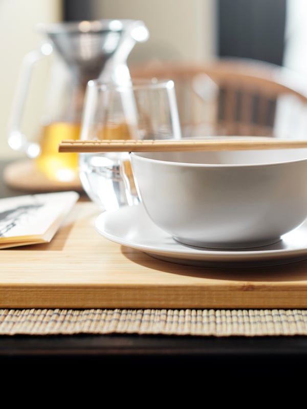 A set of bamboo TREBENT chopsticks balance on a beige DINERA bowl with a side plate underneath on a wooden board on a table.