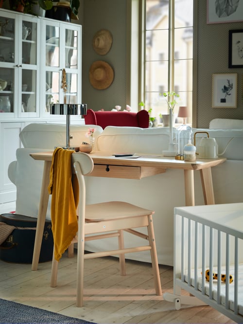 A chair and an ash veneer LISABO desk with a built-in drawer stand behind a white sofa by a white cradle in a living room.