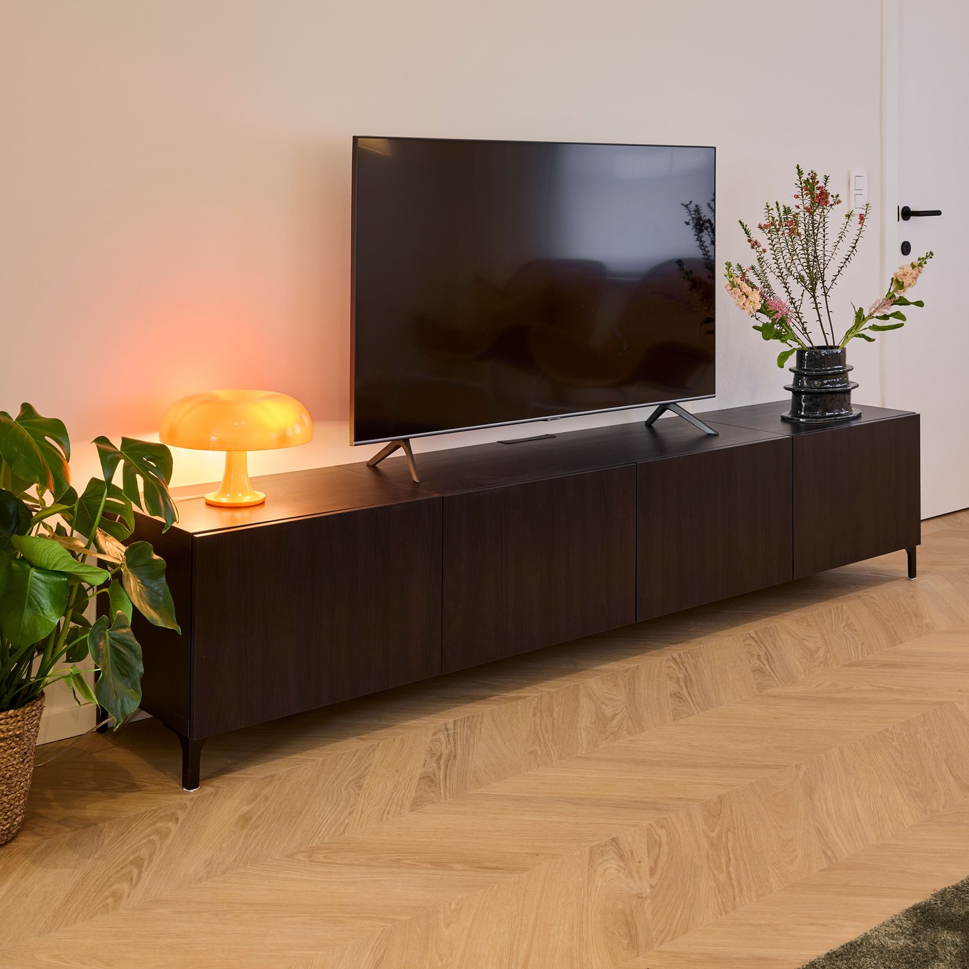 Donker tv-meubel met flatscreen, lamp en vaas naast plant op houten vloer.