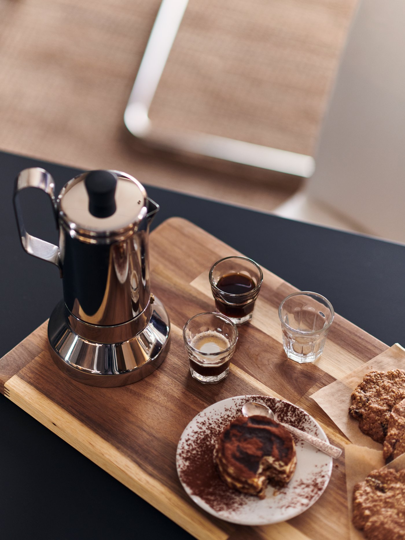 A METALLISK espresso maker, POKAL glasses, a cake and biscuits are on a SMÅÄTA chopping board on a kitchen countertop.