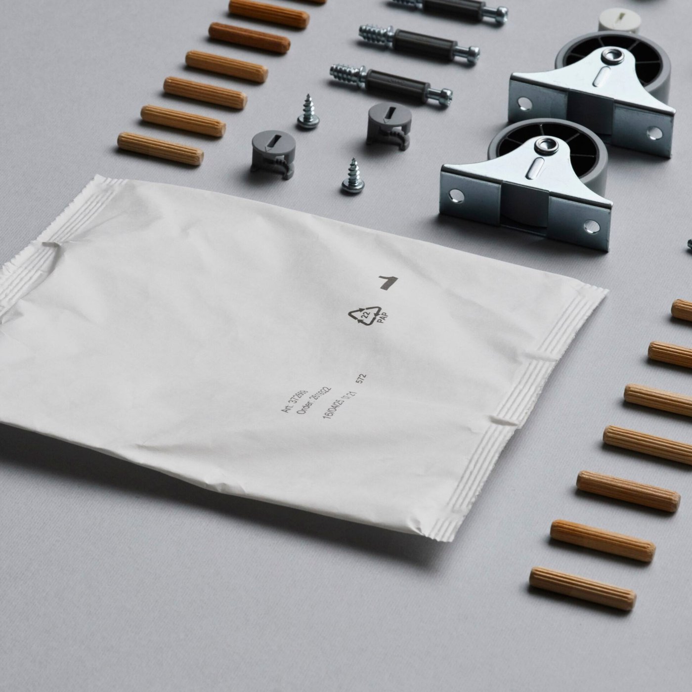 A paper-based fitting bag is next to assembly parts like screws, dowels and castors laid out neatly on a grey surface.