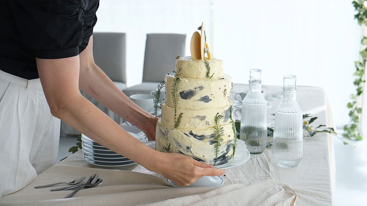 En høj moussekage på et kagefad, der står på et festligt bord med sidetallerkner og glasvandflasker på dugen.