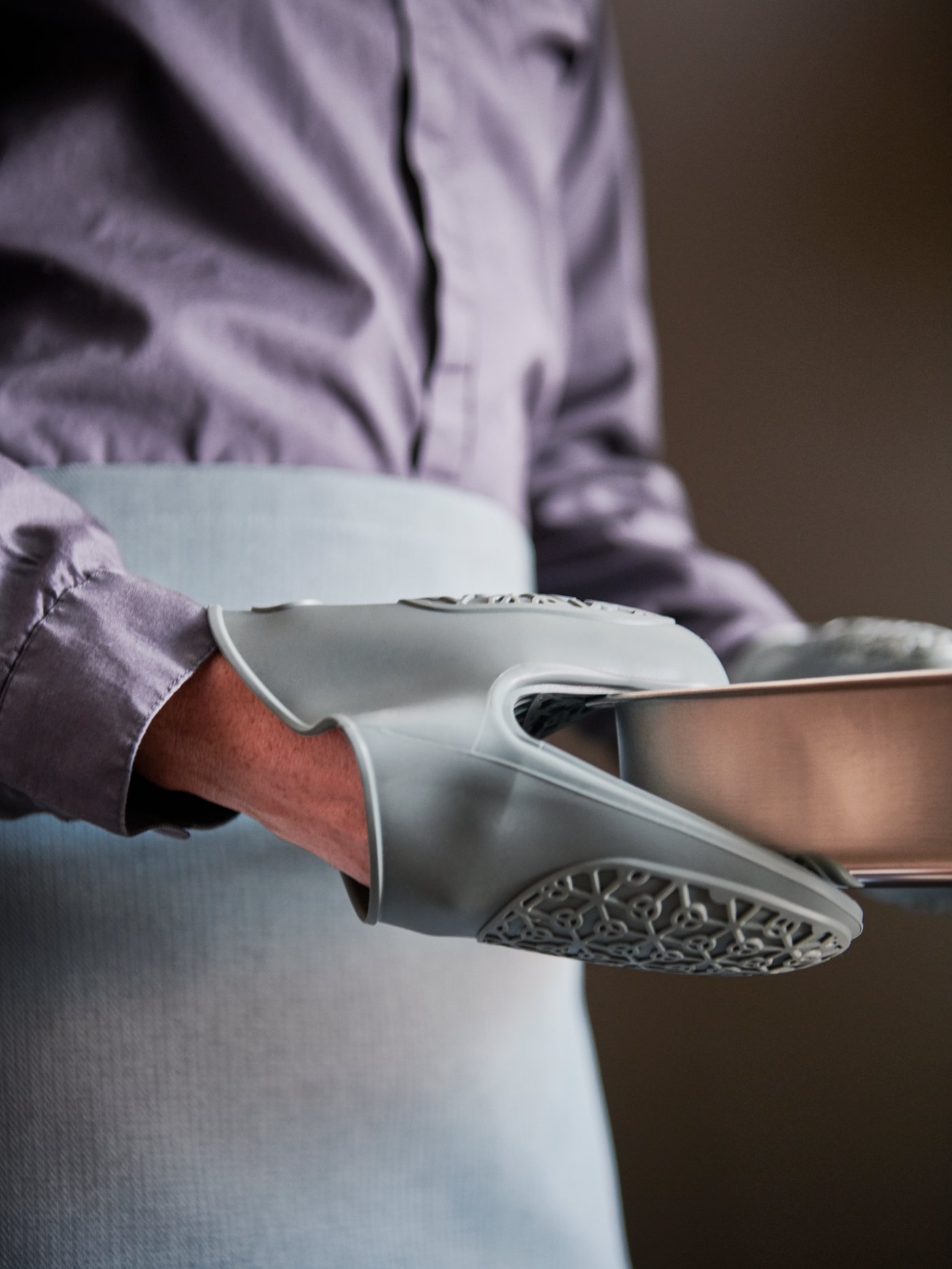 A person with a blue SANDVIVA waist apron holding a grey HEMMABAK roasting tin with a blue SANDVIVA silicone oven glove.