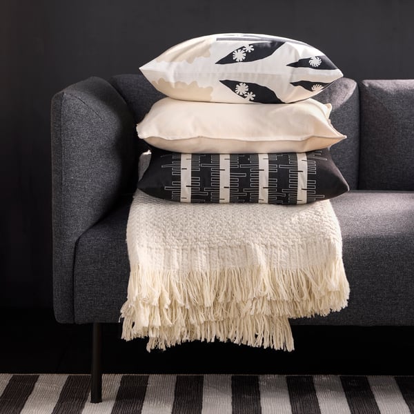 An off-white throw with three cushions stacked on it, the top one with a DRABOR cushion cover, rests on a grey sofa.