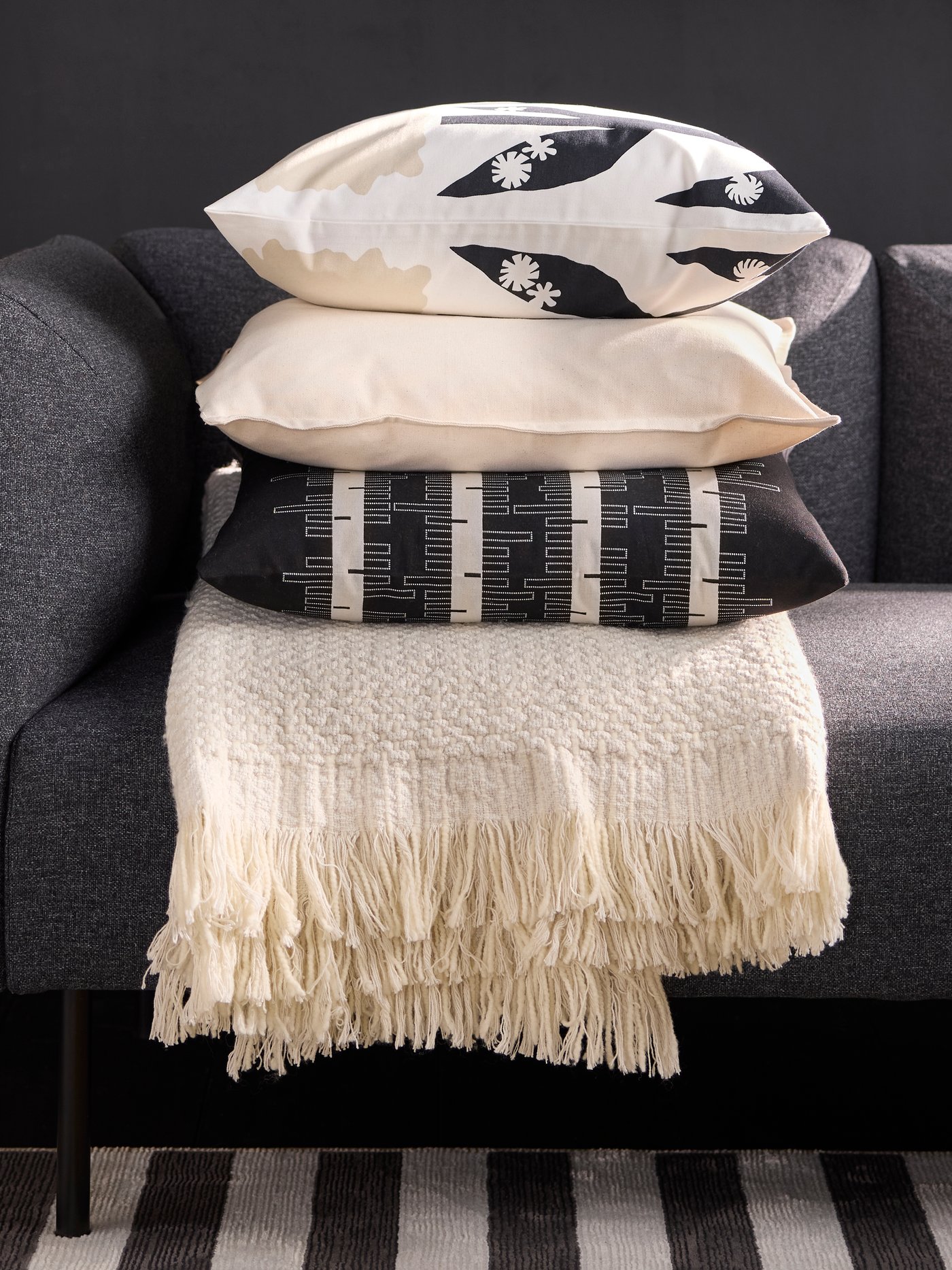 An off-white throw with three cushions stacked on it, the top one with a DRABOR cushion cover, rests on a grey sofa.