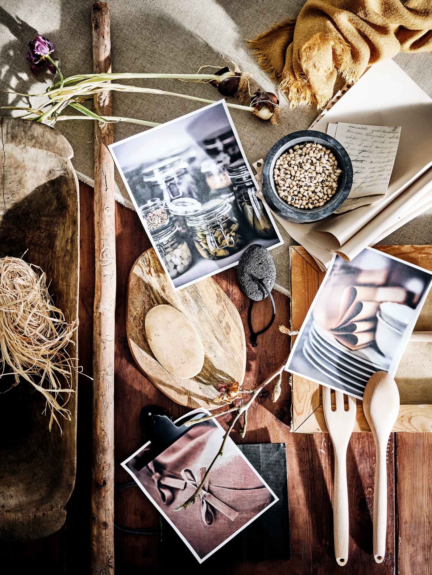 A rustic wooden table top with dried flowers, photographs, straw, a bowl, a wooden spoon, fork and chopping board on top.