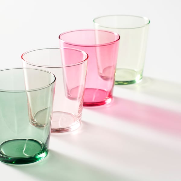Four PAPPERSBJÖRK glasses in shades of green and pink are placed in a row on a grey surface, casting colourful shadows.