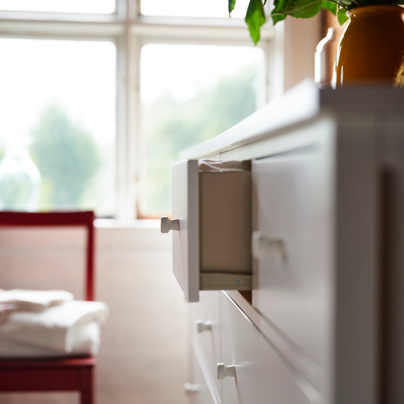 In a bedroom with timber walls, a GULLABERG chest of 8 drawers has one drawer pulled out and a plant and vase on top.