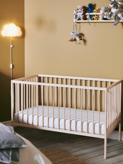 Modern baby and children room focus of a beige cot made out of solid beech and nylon fabric next to a white LED wall lamp.