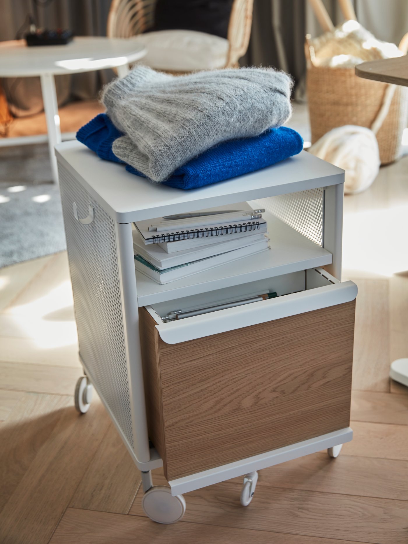 A white-mesh BEKANT storage unit on castors standing on a wooden floor, holding paperwork and knitted garments on top.