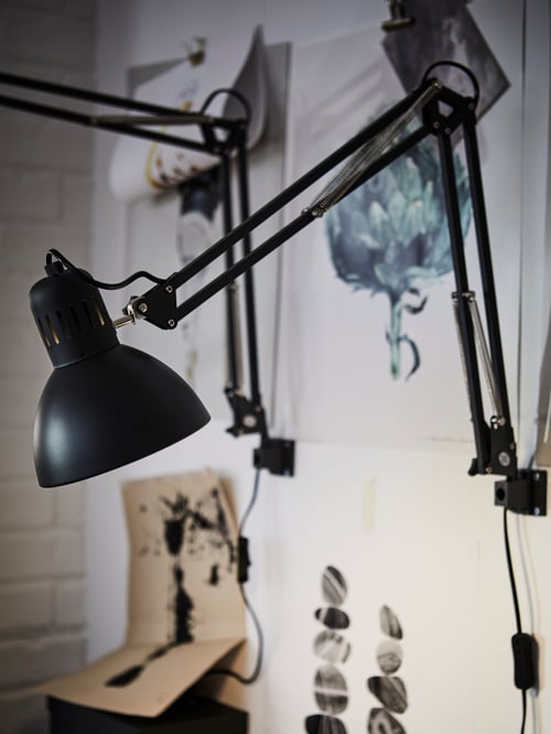 Eine Nahaufnahme einer TERTIAL Arbeitsleuchte in Dunkelgrau an der Wand befestigt. Im Hintergrund sind Illustrationen zu sehen.