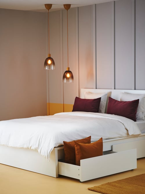 Bedroom with two pendant lamps mounted above a high bed frame with pillows, with an open storage box with pillows underneath.