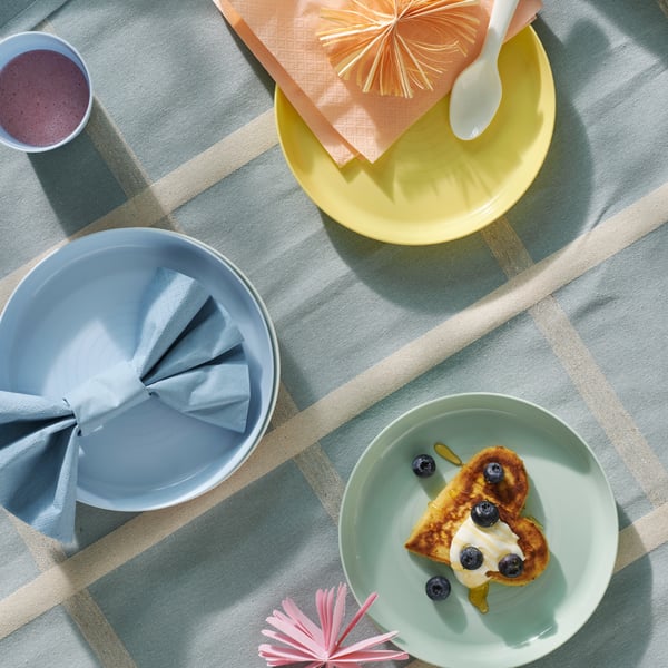 A birds-eye view of pastel coloured KALAS children’s plates on a light blue check table cloth. One of them has food on it.