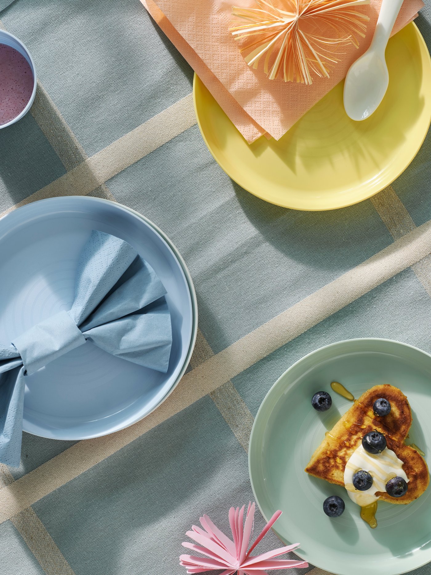 A birds-eye view of pastel coloured KALAS children’s plates on a light blue check table cloth. One of them has food on it.