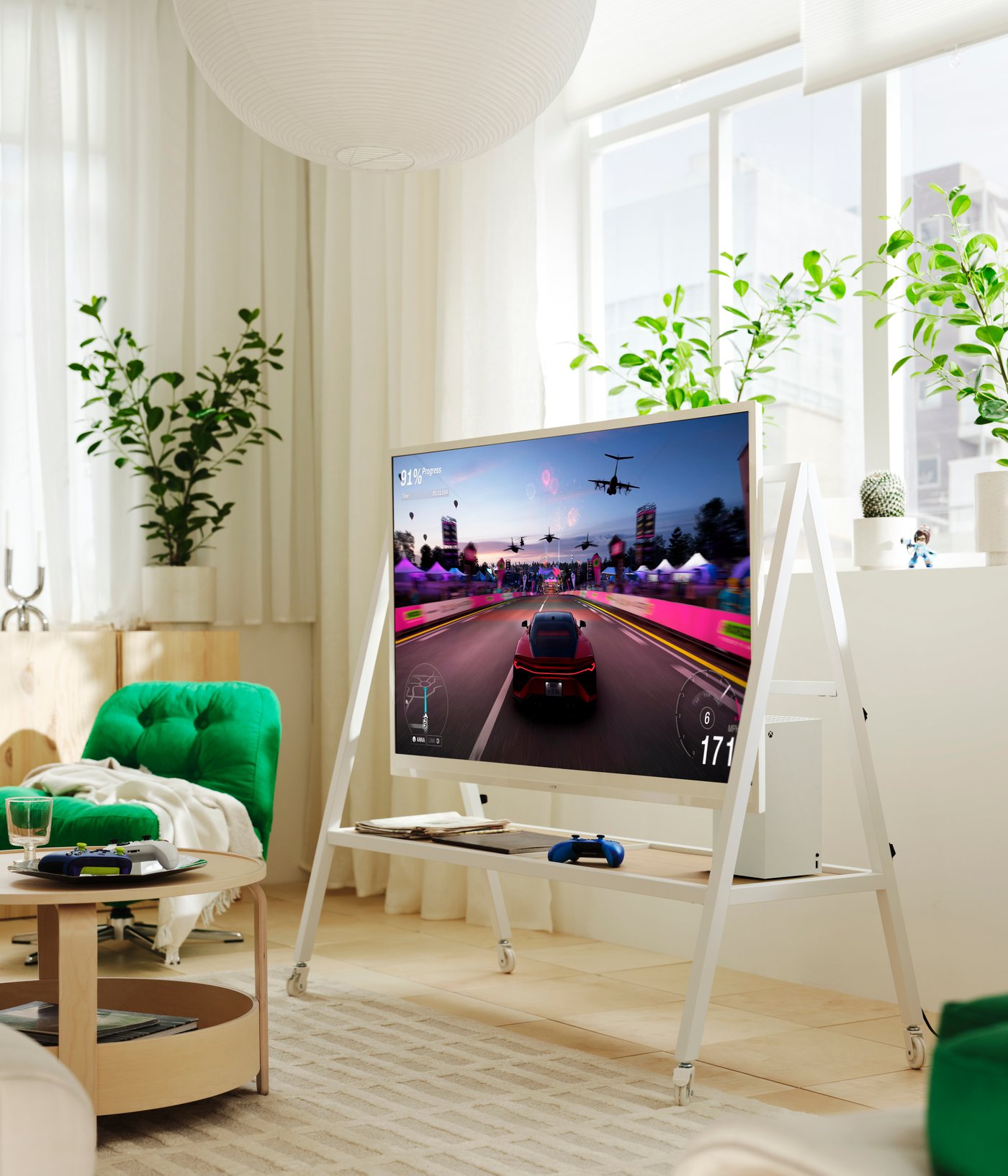 Ein weißer YXSTABY Gaming-TV-Schrank auf Rollen steht vor einem Fenster in einem hellen Wohnzimmer. Auf dem Fernseher ist ein Rennspiel zu sehen.