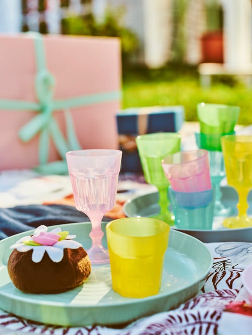 A picnic blanket set with DUKTIG multicolour glasses, KALAS mixed-colour plates, toy food and wrapped gifts.