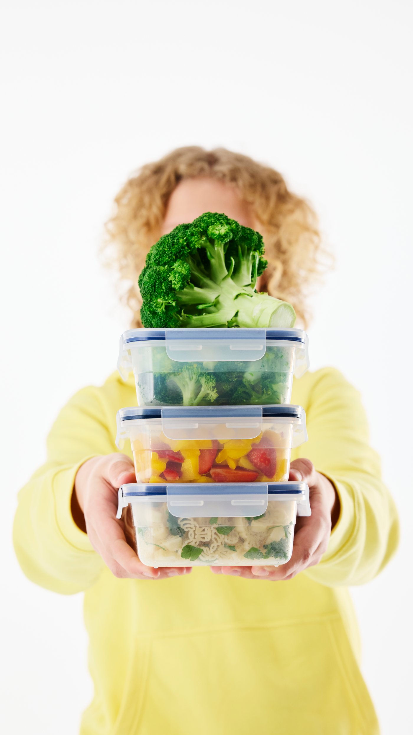 Žena v žluté mikině drží v natažených rukou stoh plastových dóz s modrými víčky naplněných zeleninou, nahoře vykukuje brokolice.