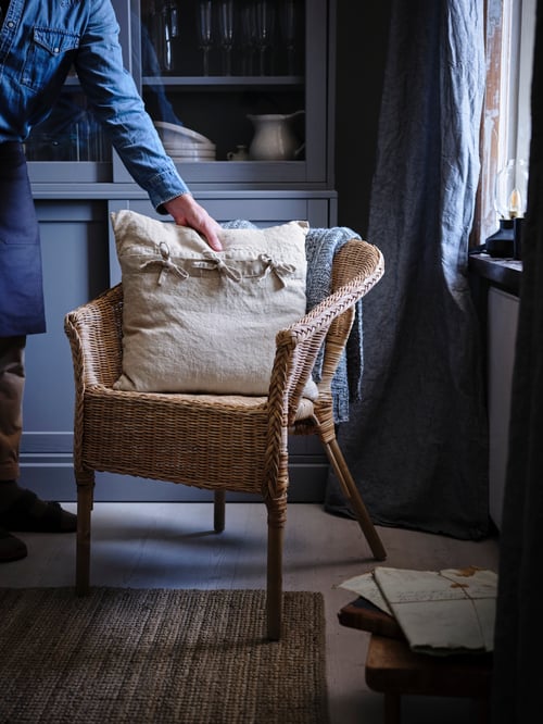 Salon avec une chaise beige, une housse de coussin, un tapis, un meuble de rangement avec portes vitrées, des rideaux, une personne, des lunettes.