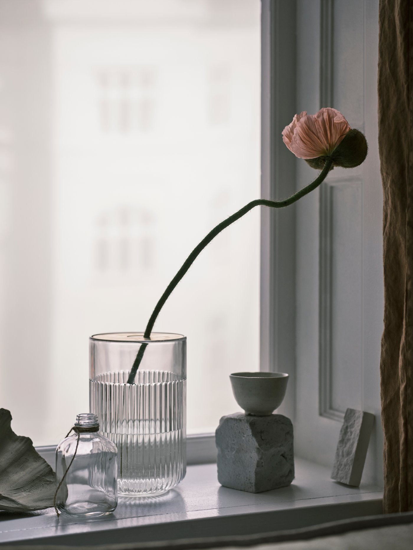 A long-stemmed flower in a GRADVIS glass vase on display in a windowsill, next to a glass bottle and a small grey bowl.