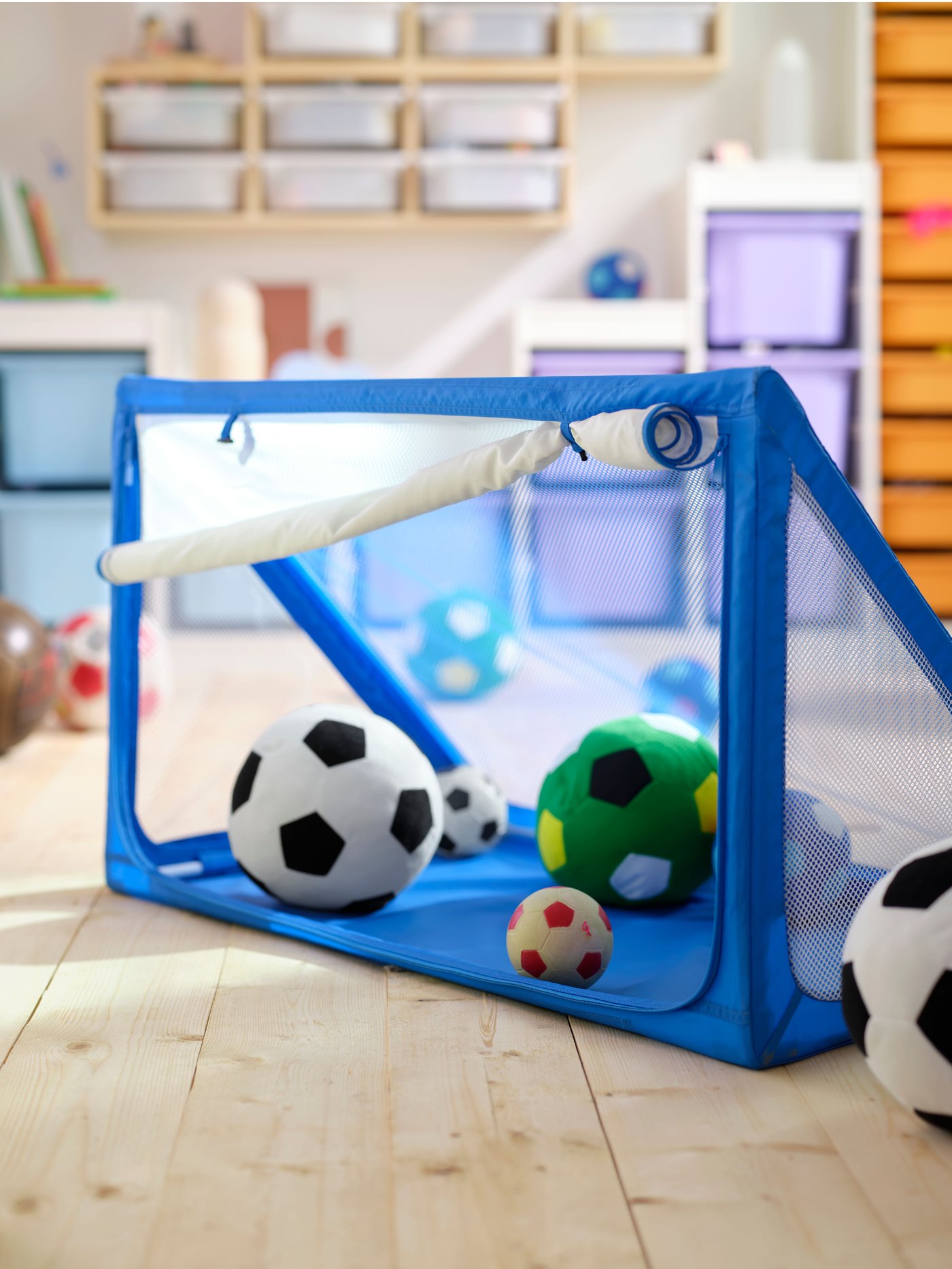 A SPORTSLIG ball storage/goal is placed on the floor in a room. There are several SPARKA soft toy footballs in and around it.