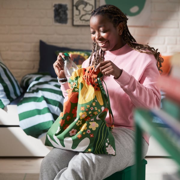 A person in a bedroom sits on a green stool and puts a bottle into a green CAFEBITRÄDE drawstring bag with a floral pattern.