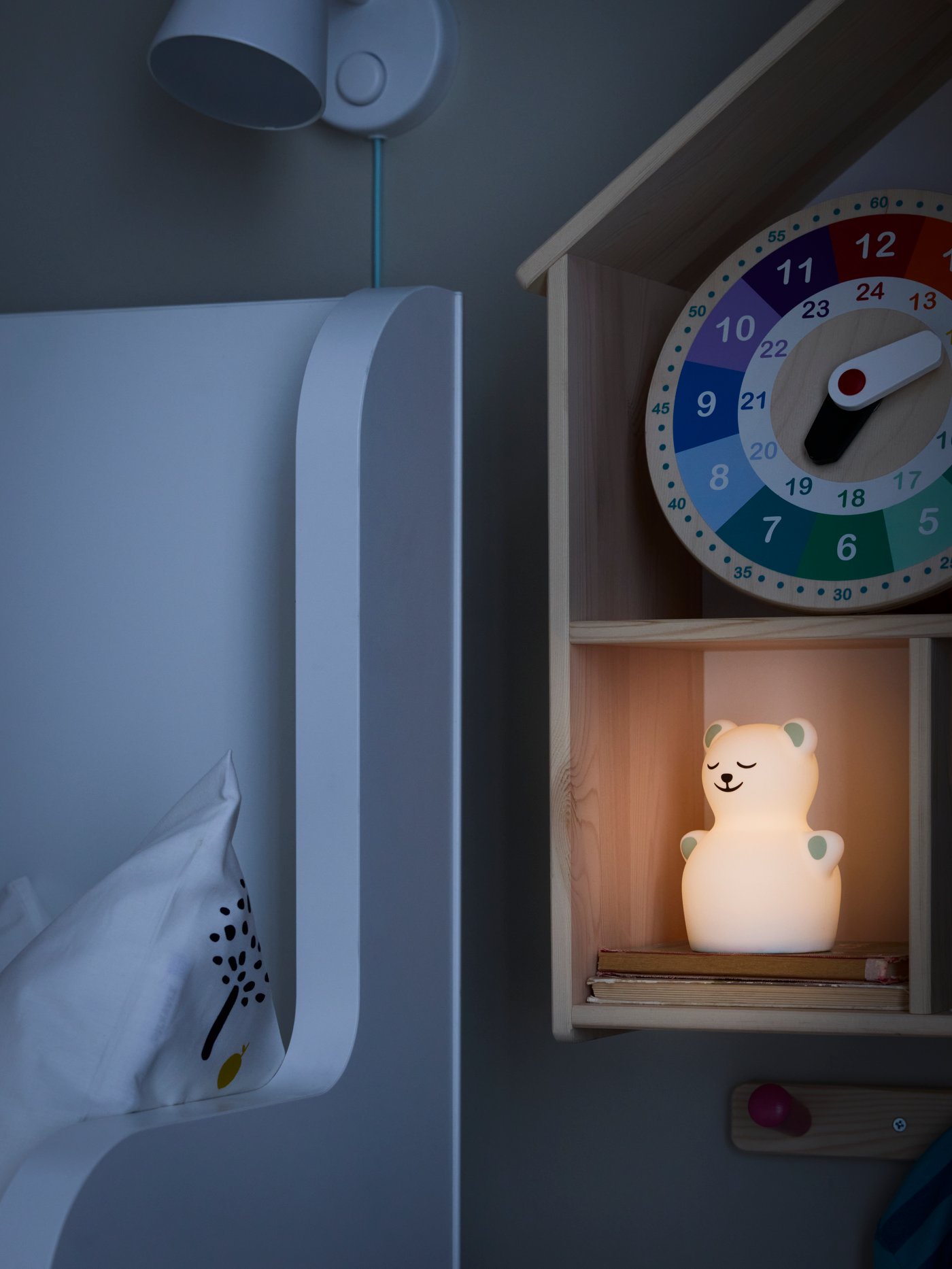 Una lámpara de noche TÖVÄDER encendida y un reloj de madera multicolor UNDERHÅLLA en una casa de muñecas de pared, junto a una cama.