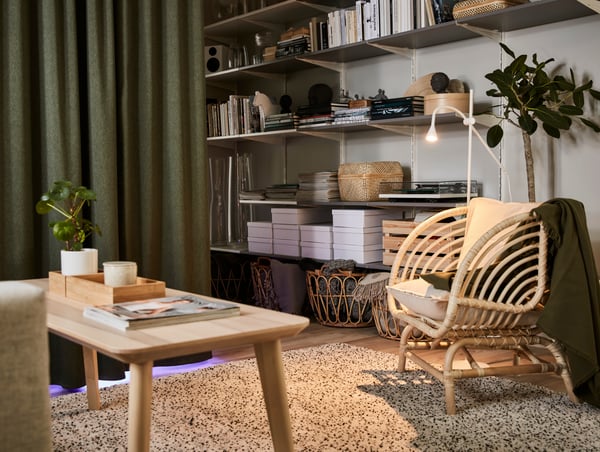 Ein Wohnzimmer mit einem Couchtisch in Eschenfurnier, einem Sessel in Rattan, einen hochflorigen Teppich und grünen raumaufteilenden Verdunklungsgardinen