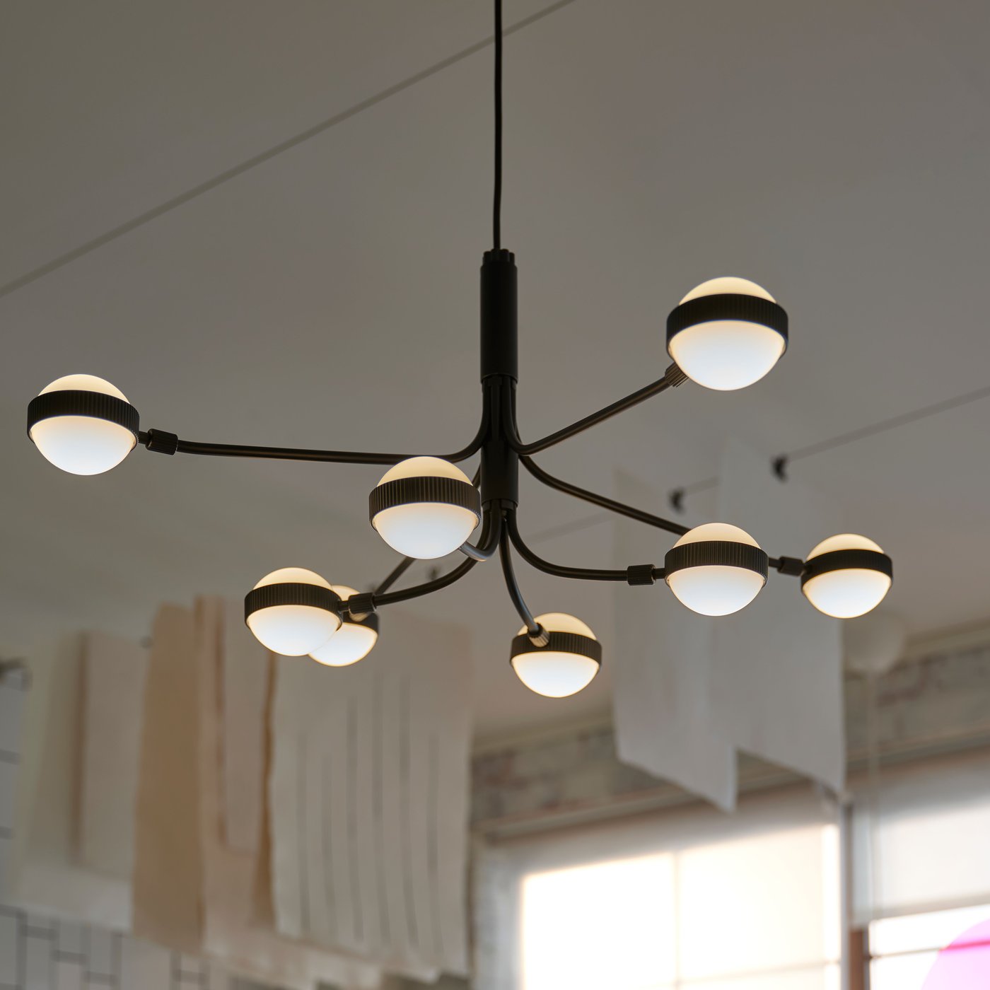 A black/white 8-armed STORSLINGA LED chandelier hangs from a ceiling in an industrial-style room with large windows.