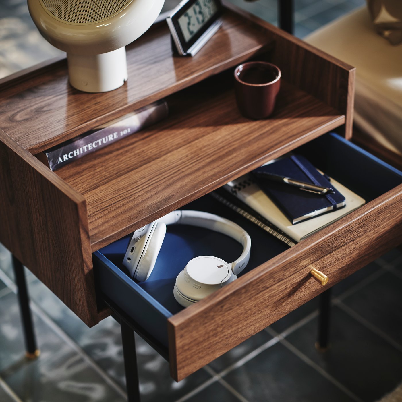 Close-up of RÅDMANSÖ bedside table in a walnut colour with some knick-knacks and BLOMPRAKT Bluetooth speaker lamp on top.