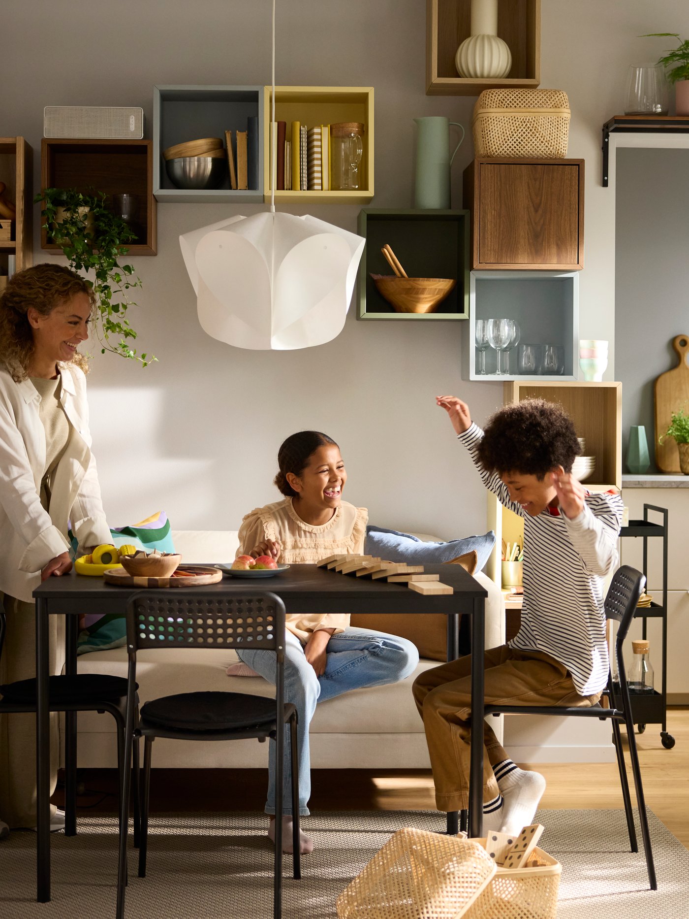 A mum and two children in a sunlit dining area of a SANDSBERG table flanked by ADDE chairs, a MARIUS stool and a sofa.