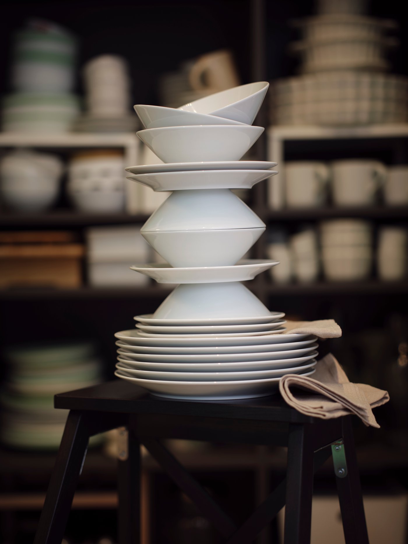 Weißes Geschirr zu einem hohen, verspielten Turm gestapelt auf einem Hocker; daneben ein beigefarbenes Tuch, im Hintergrund Regale mit weiteren Tellern und Tassen – ordentlich, alltagsbereit.