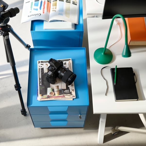 Aerial shot with bright blue storage cabinet on one side and corner of white desk with green angle poise lamp on the other.