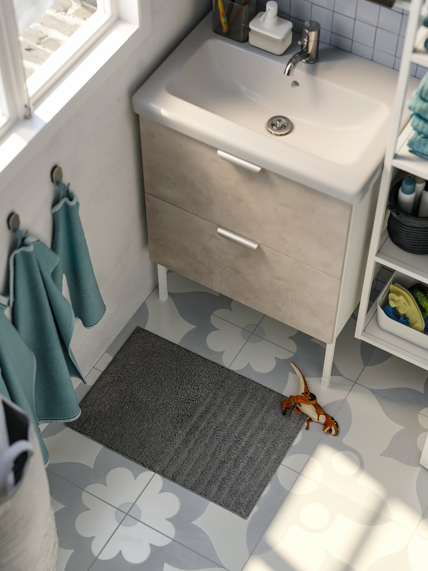 Un tapis de bain gris sur lequel se trouve un dinosaure jouet; des serviettes bleu clair suspendues à des crochets muraux, un lavabo blanc.