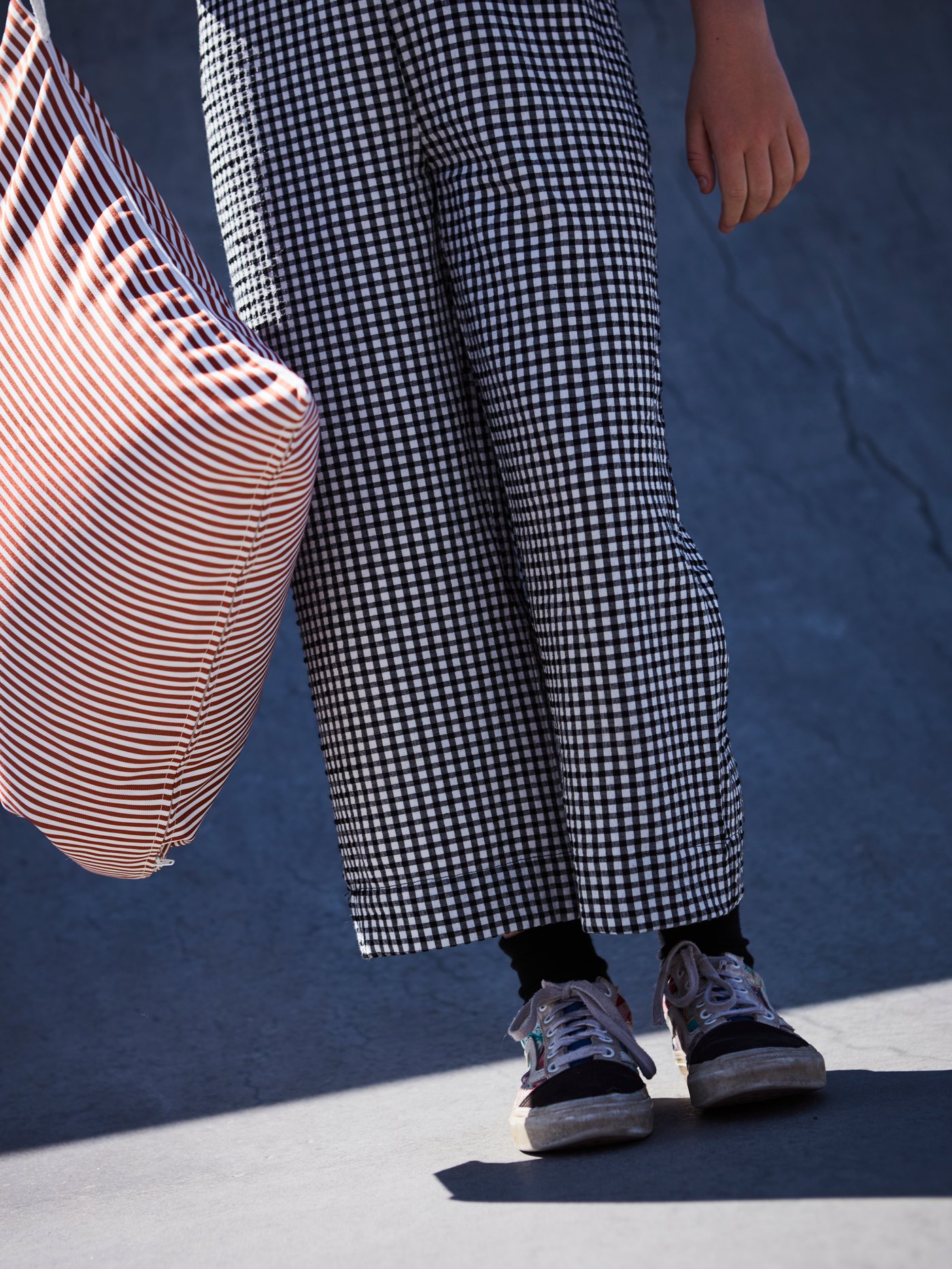 A person wearing black and white checkered pants holding a GULLBERGSÖ cushion in red and white stripes in a sunny place.