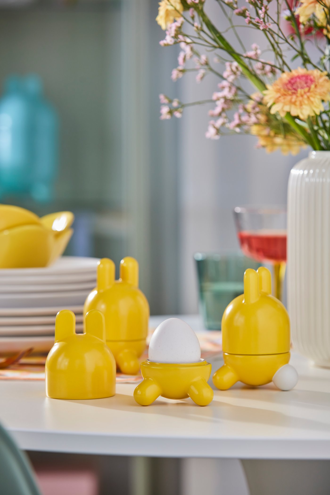Three yellow TJÄRLEK egg cups are placed on a white tabletop. The cup in the middle has no lid on, and there is an egg in it.