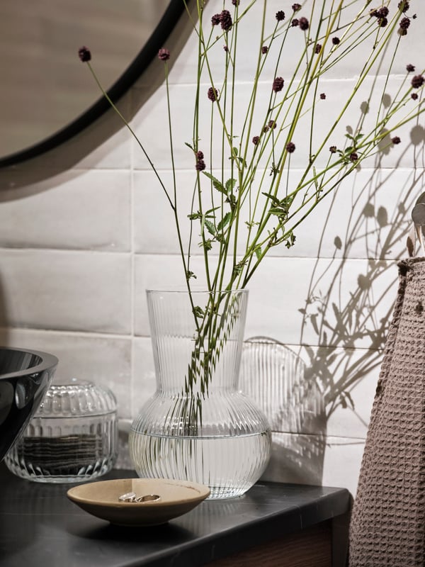 En PÅDRAG i klarglas bredvid en behållare på en bänkskiva i TOLKEN i svart marmoreffekt och en VALLASÅN handduk.