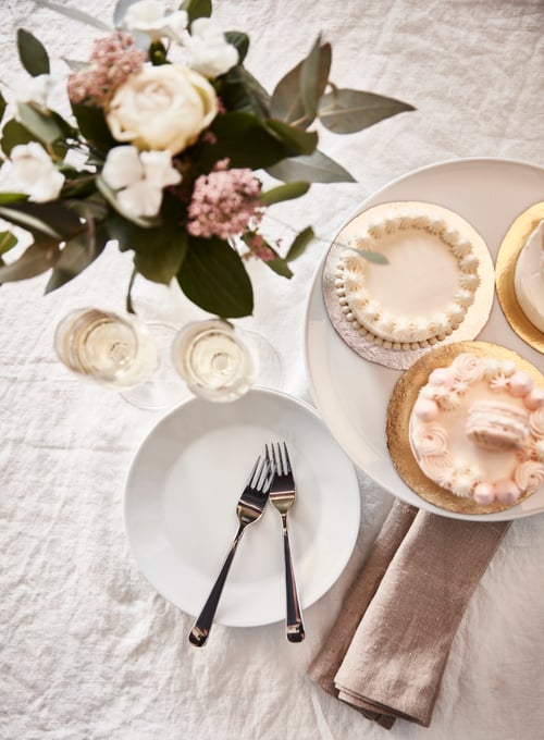 Blick von oben auf ein Tischarrangement mit zwei DRAGON Kuchen-/Dessertgabeln, einem IKEA 365+ Teller, einem UPPLAGA Kuchenteller mit Miniaturtorten und zwei STORSINT Sektgläsern.