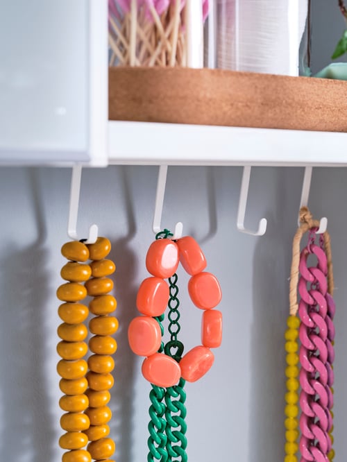 Hooks with necklaces in different colours, hanging from a white ENHET frame, with bathroom items on the shelf above.