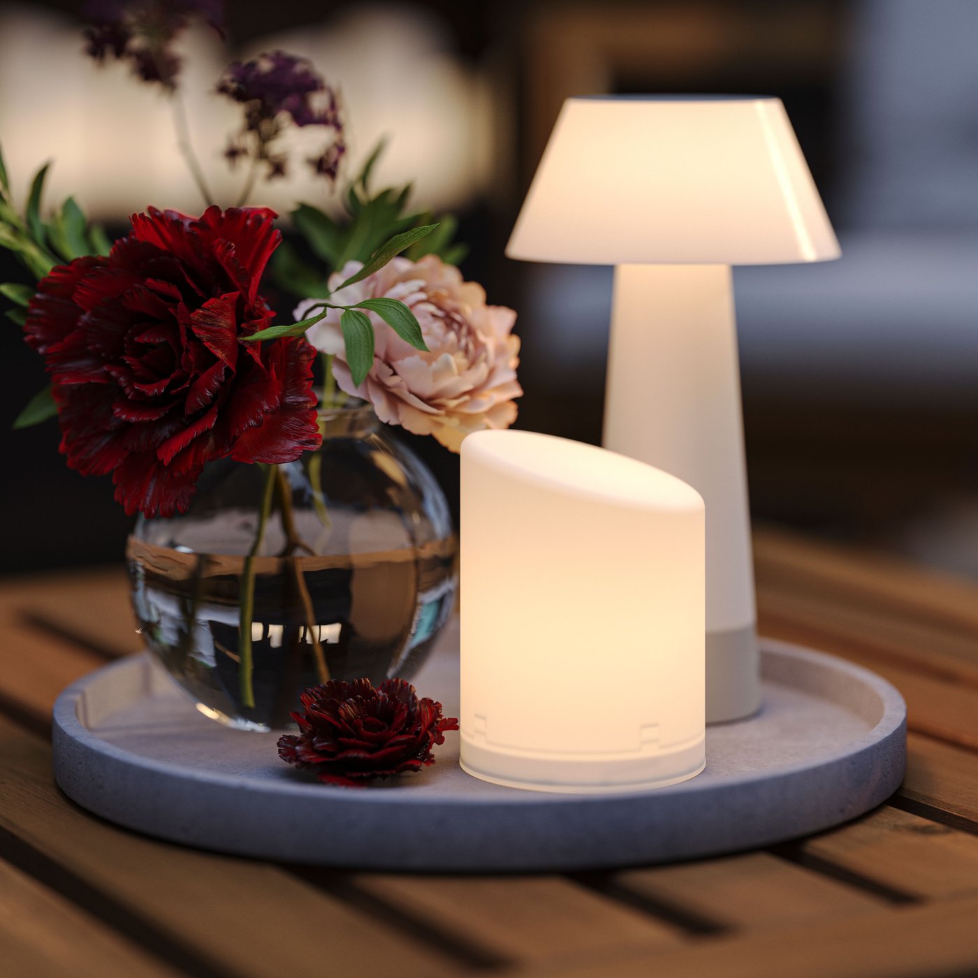 A lit in/outdoor ÄDELLÖVSKOG LED block candle and a battery-operated table lamp on a light grey saucer on a wooden table.