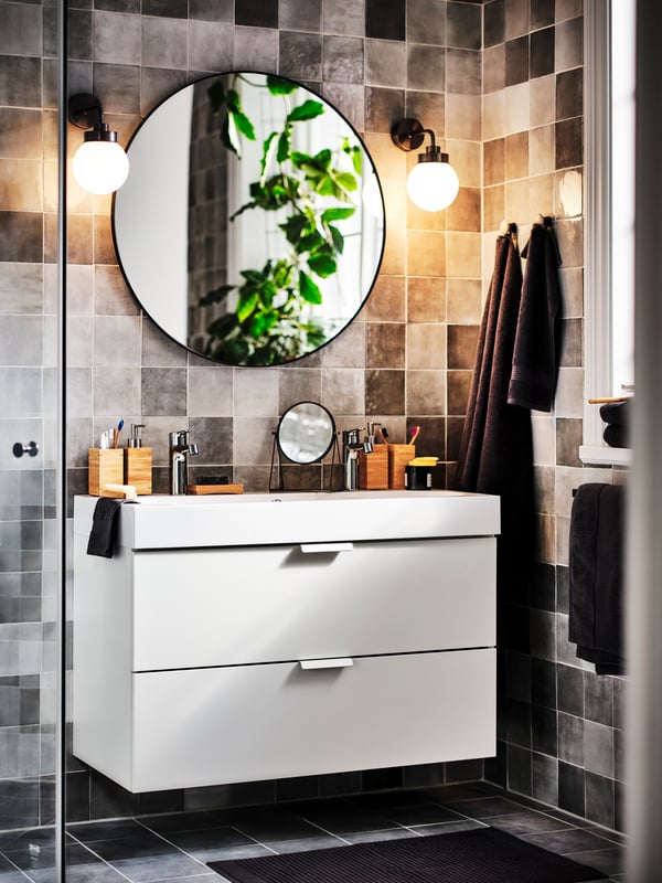 Two lit black FRIHULT wall lamps on either side of a black LINDBYN mirror above a white GODMORGON wash-stand with 2 drawers.