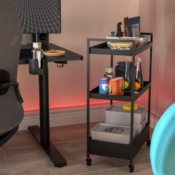 A NISSAFORS trolley in black is next to a gaming desk. The trolley has snacks, beverages and storage boxes on its shelves.