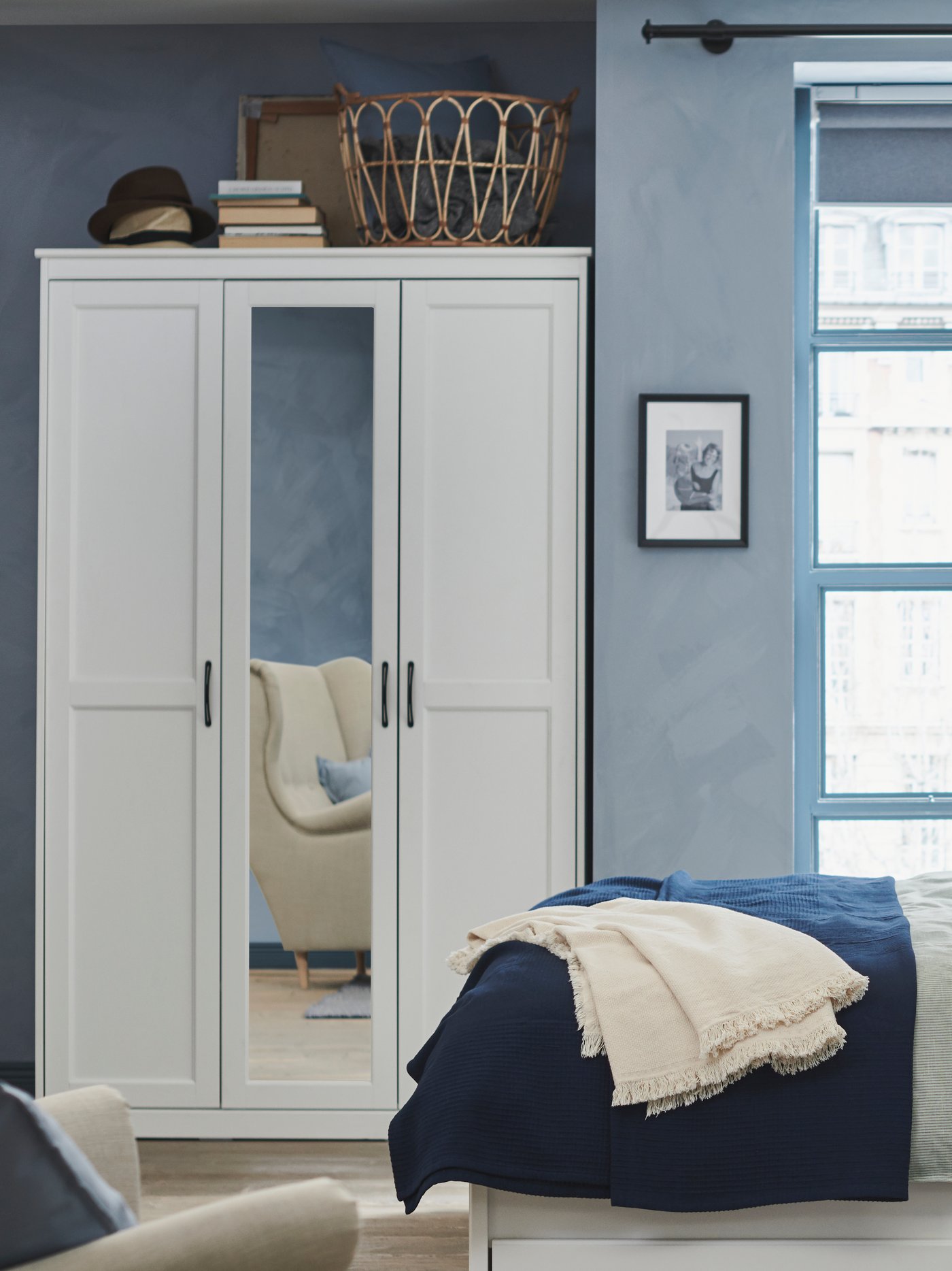 Ljust sovrum med vit garderob med spegeldörr, blå väggar, bäddad säng med blått överkast och beige pläd, samt korg och böcker ovanpå garderoben.
