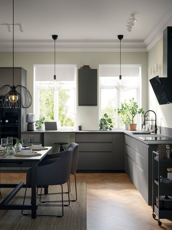 Part of a large kitchen with smooth dark grey VOXTORP fronts has two windows and a dining area and three black pendant lamps.