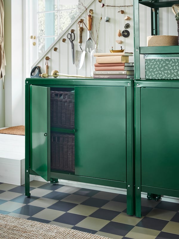 Garden tools on hooks hang on the side of the stairs behind green KOLBJÖRN cabinets with books and a box on top.