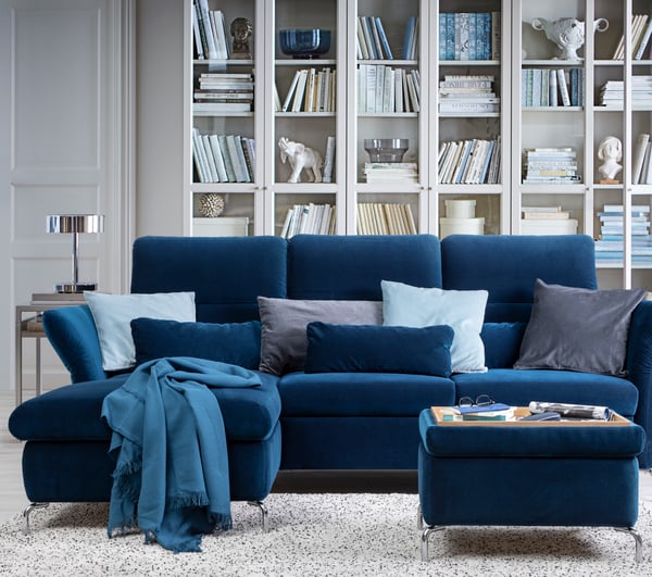 Ein Ecksofa mit einem dunkelblauen Samtbezug steht vor einer Bücherregalwand mit Vitrinentüren. 